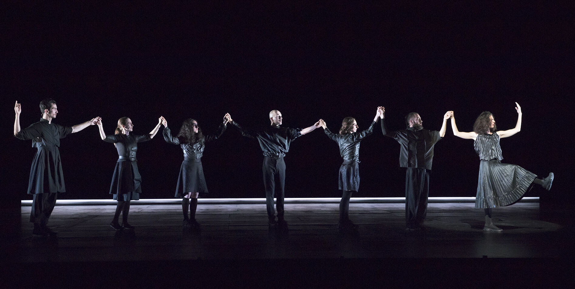 ANΩNYMO: seven dancers in dark formal clothing stand in a wide line across a black stage, holding hands with arms raised on either end.