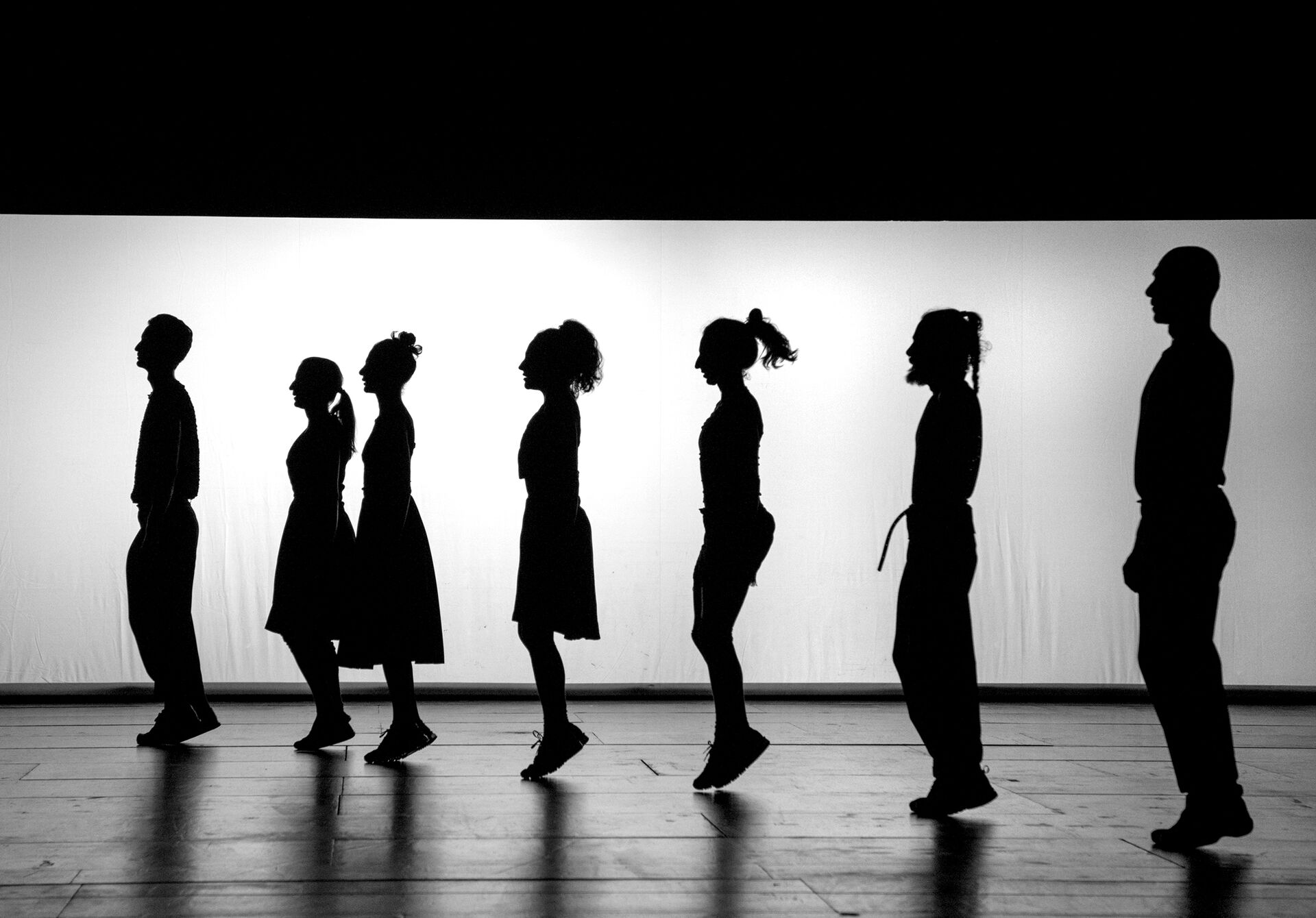ANΩNYMO: seven dancers shown in silhouette, walking in profile from left to right across a brightly back-lit white screen.