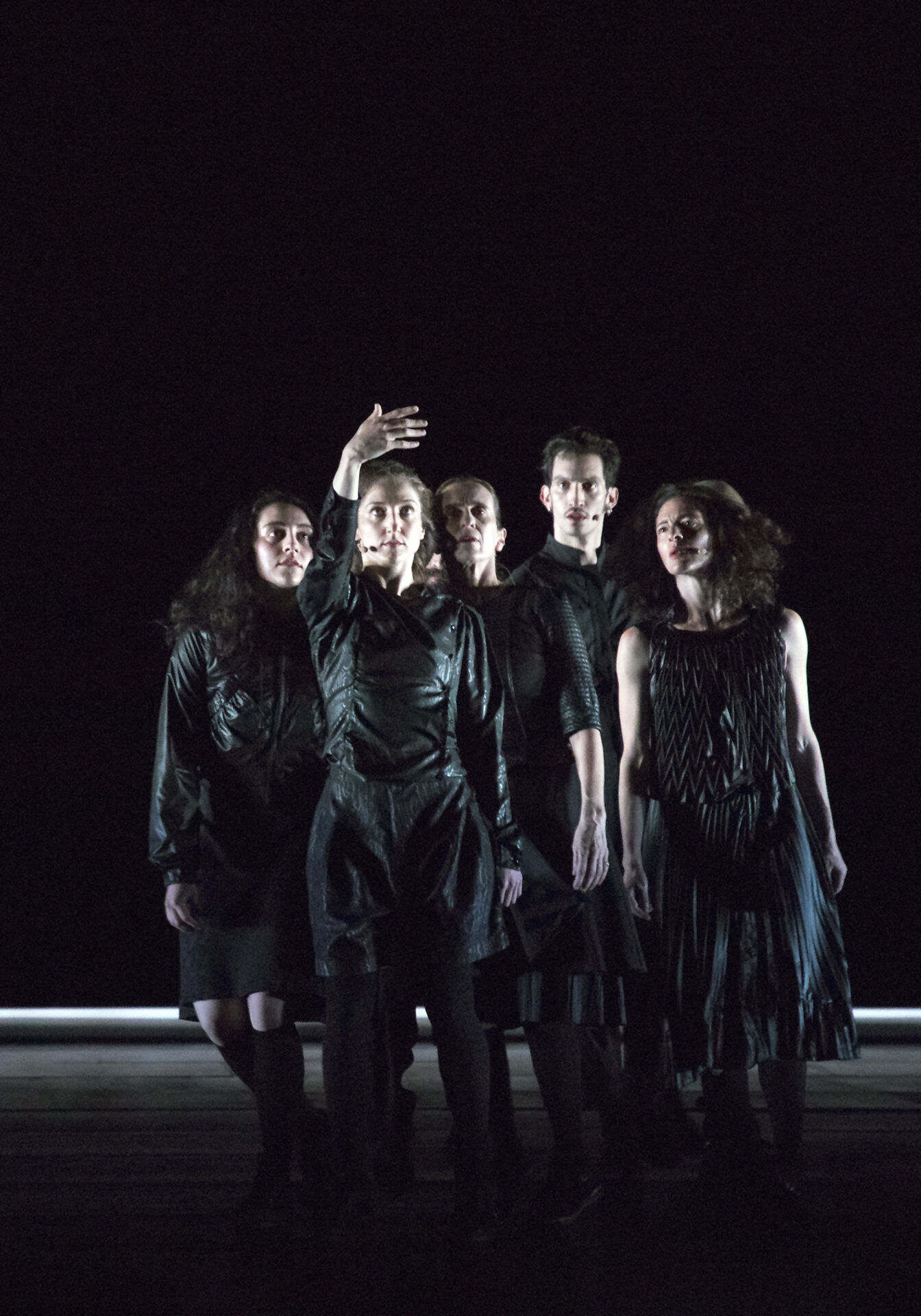 ANΩNYMO: five dancers in dark clothes stand close together on a dimly lit stage; a woman at the front raises one arm high while the others gather behind her.
