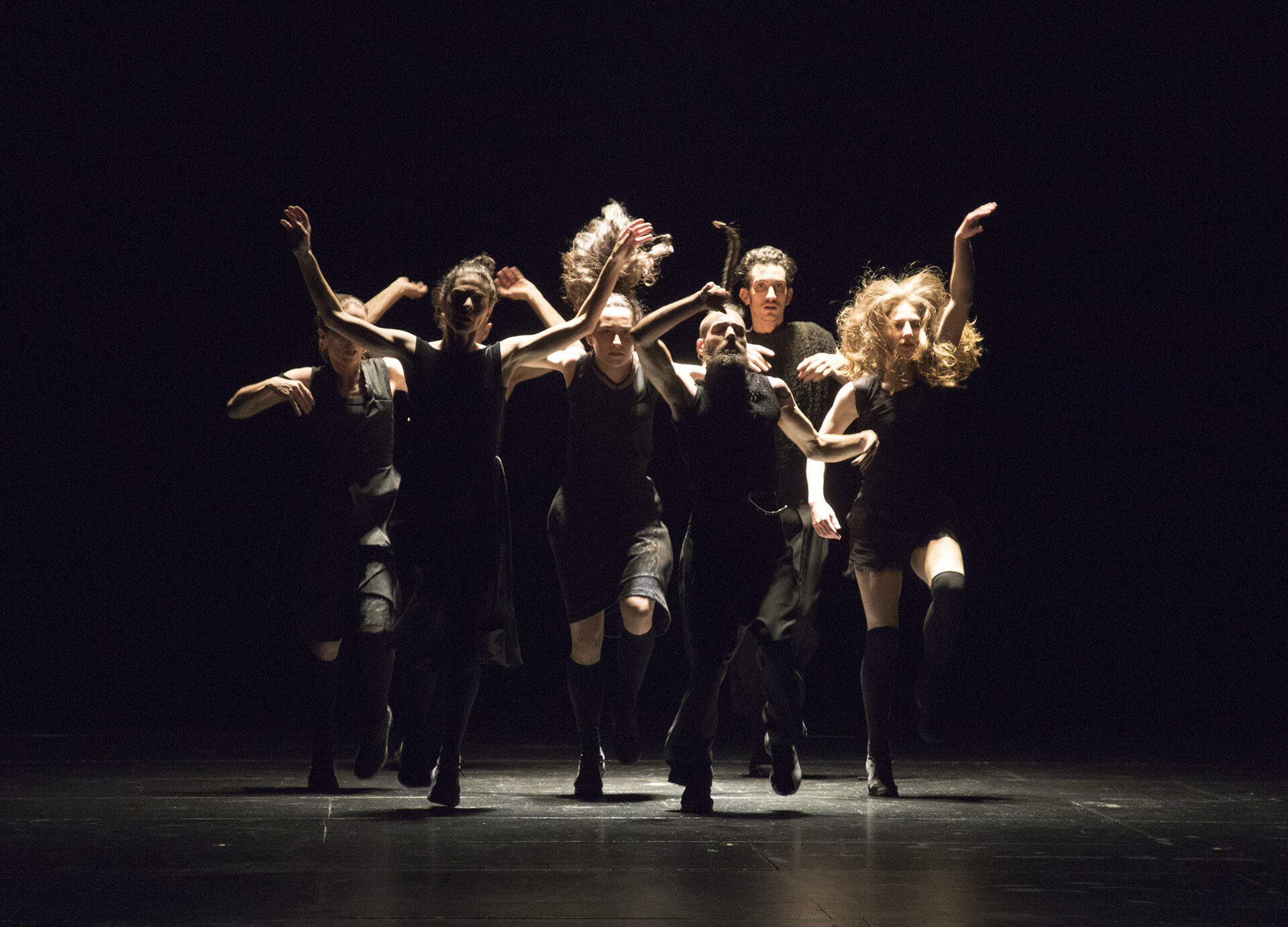 ANΩNYMO: a group of dancers in black clothing move forward together in a tight cluster, arms raised and hair flying, lit from behind on a dark stage.