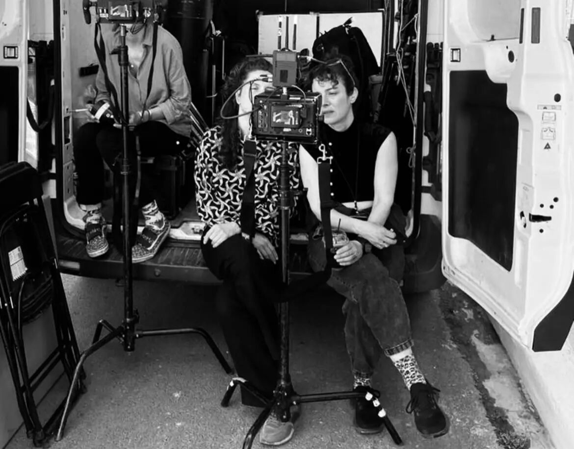 Black-and-white behind-the-scenes photo: co-directors Elke Margarete Lehrenkrauss and Youla Boudali sit at the open back of a van around a tripod-mounted cinema camera, a crew member behind them.