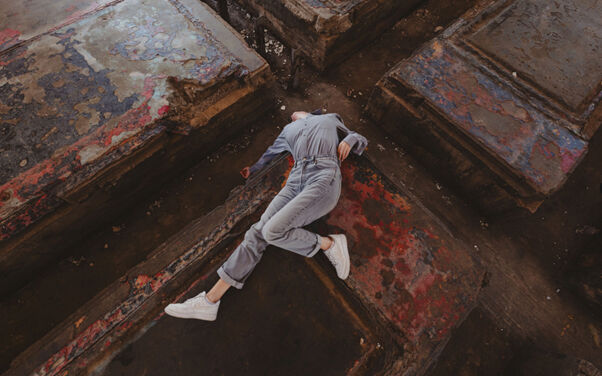TELOS: aerial view of Ermira Goro lying face-down across a dark, paint-stained wooden surface in a grey jumpsuit and white sneakers, one leg extended.