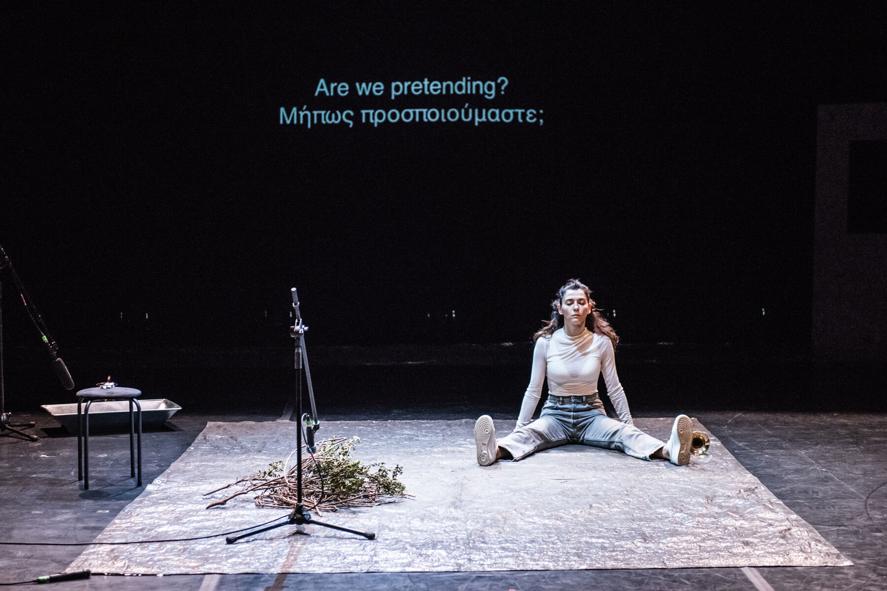 TELOS: Ermira Goro seated in a wide split on a textured grey mat at centre stage; behind her, projected text reads ‘Are we pretending? Μήπως προσποιούμαστε;’; a microphone stand and dry branches sit nearby.