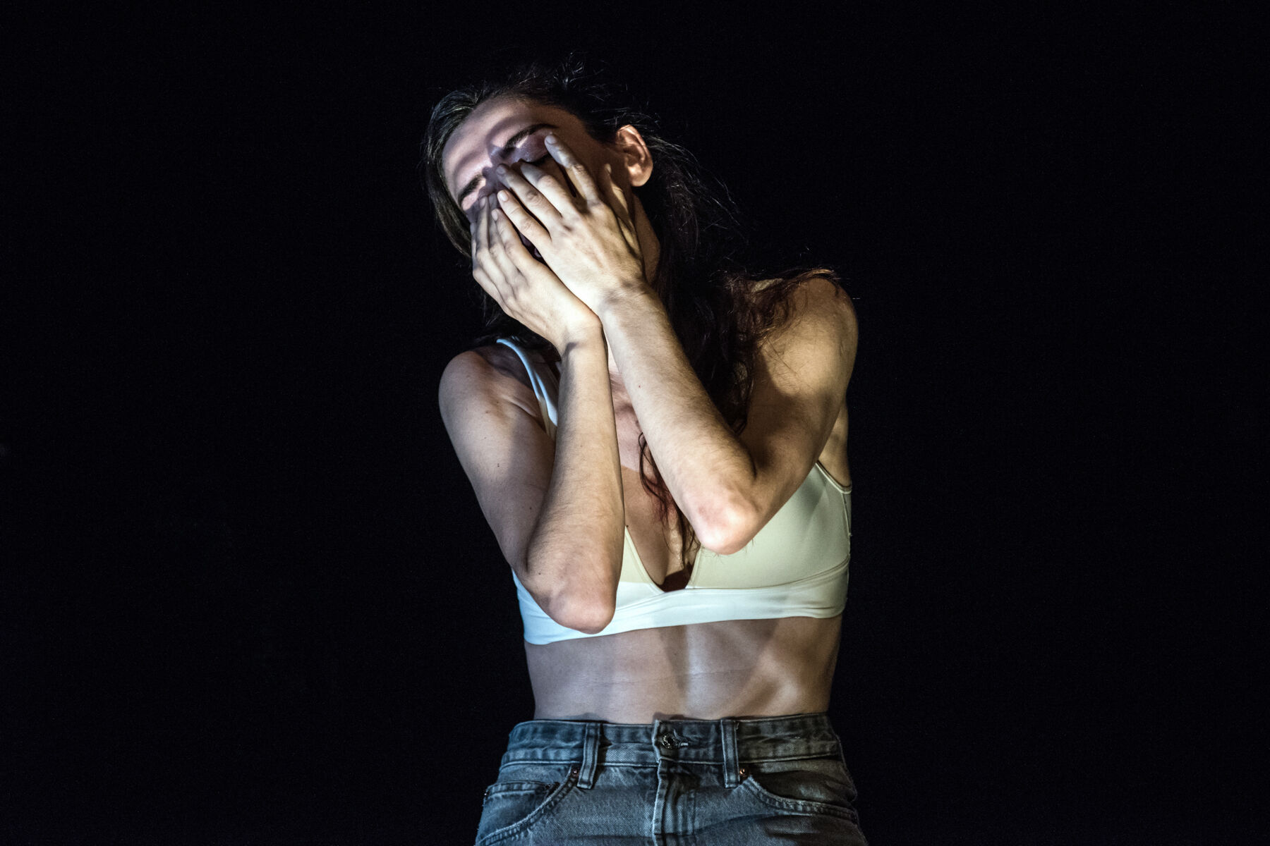 TELOS: close-up of Ermira Goro pressing both hands to her face, hair falling over her arms; she wears a cream sports bra and jeans against a black background.