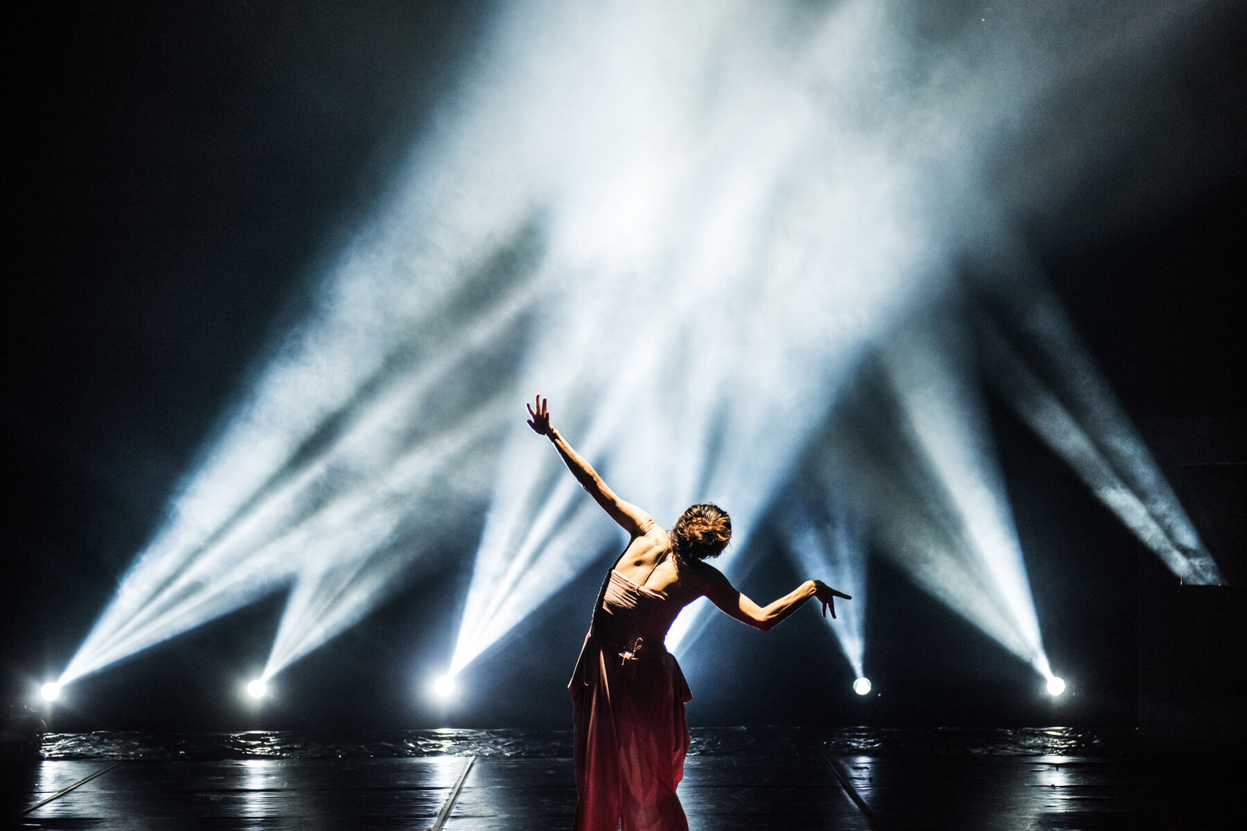 TELOS: silhouette of Ermira Goro in a dark dress with one arm raised, photographed against a stage backlit by a fan of white light beams cutting through haze.