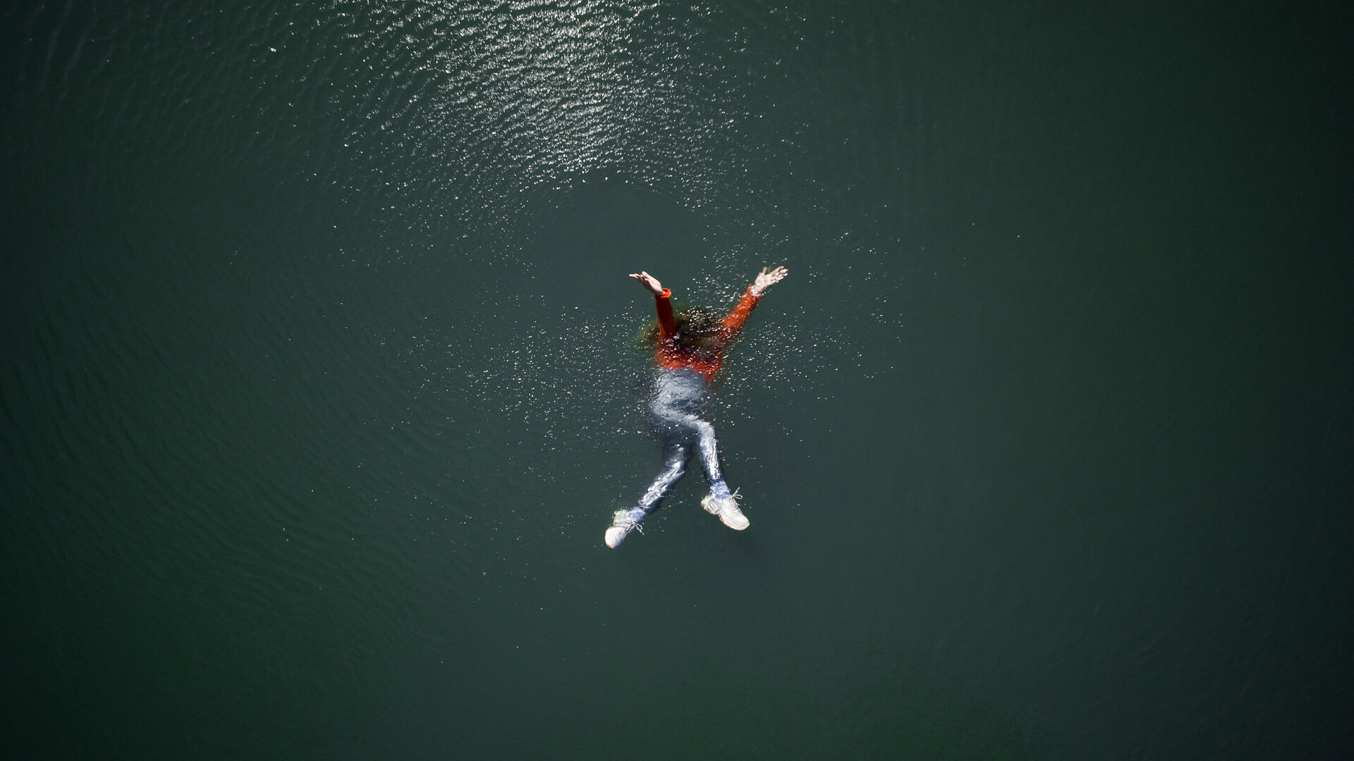 LAPSE installation still: a woman in a red top and jeans floats face-up with arms spread on the dark green surface of a wide river, photographed from directly above by Lars Jan.