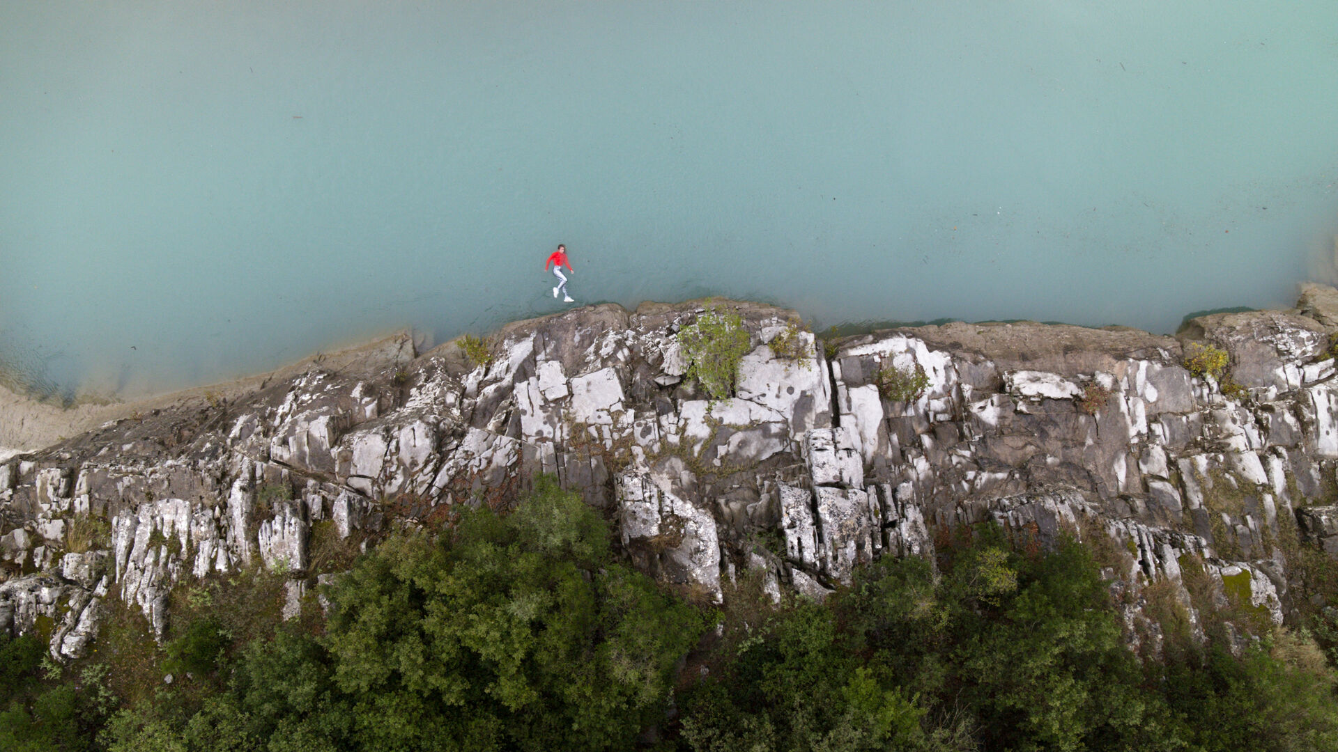 LAPSE installation still: aerial view of the same woman in red walking along a rocky white cliff edge above pale turquoise water, with dense green vegetation below, by Lars Jan.