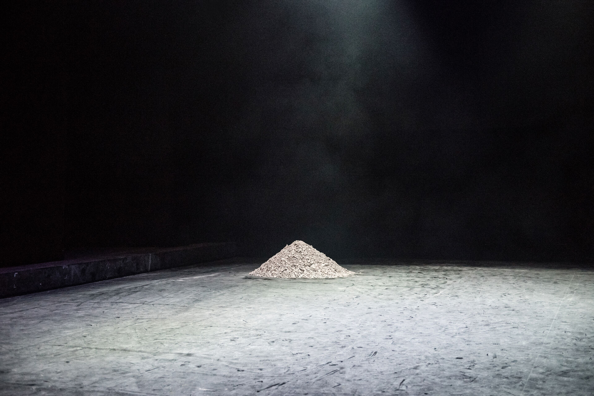 MINTATI stage shot: a single conical pile of fine grey earth at centre stage under one overhead spotlight on a dark, dust-covered floor. Photo by Karol Jarek.
