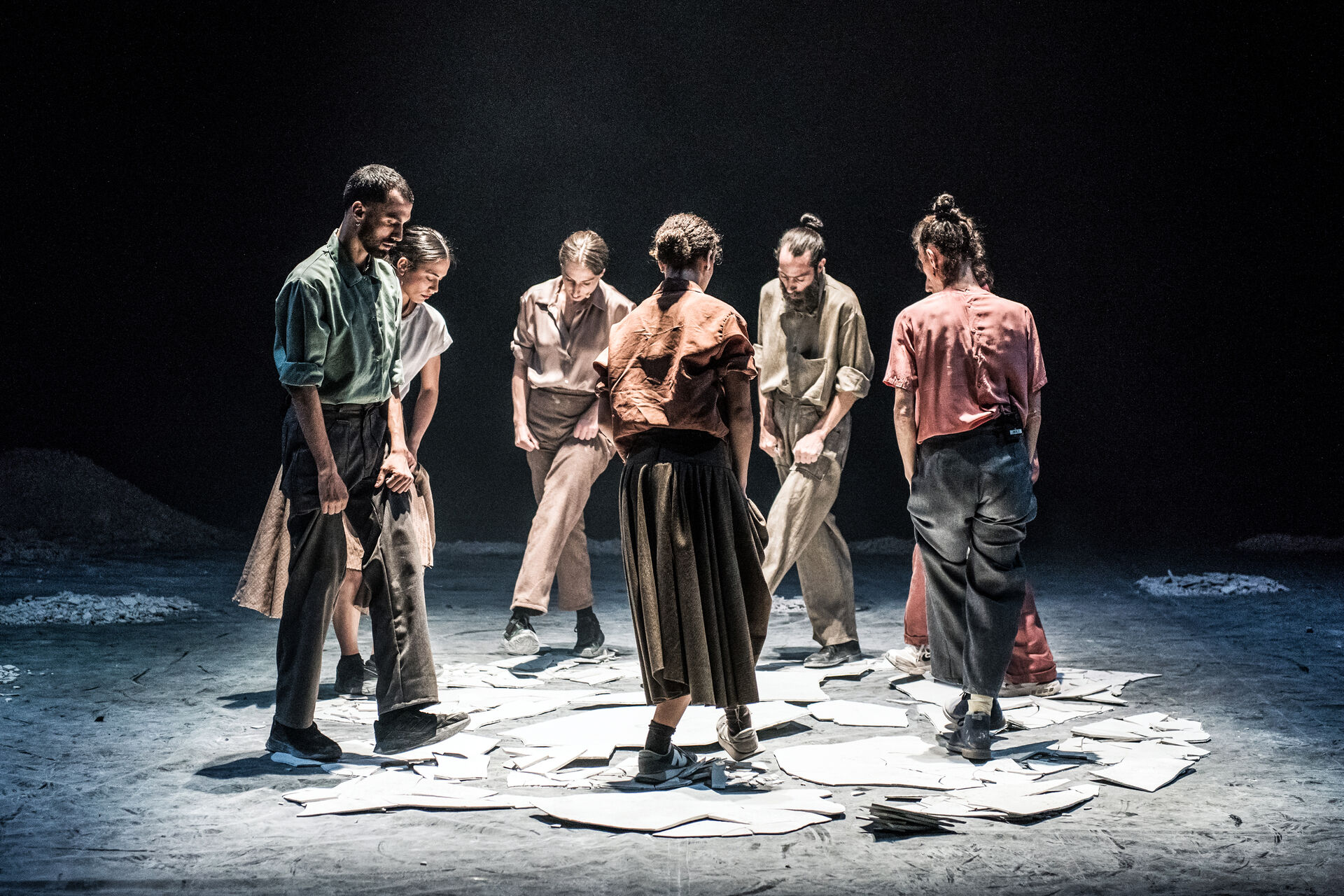 MINTATI: a circle of seven dancers in earth-toned costumes converge inward over scattered torn paper sheets on a dust-covered stage. Photo by Karol Jarek.