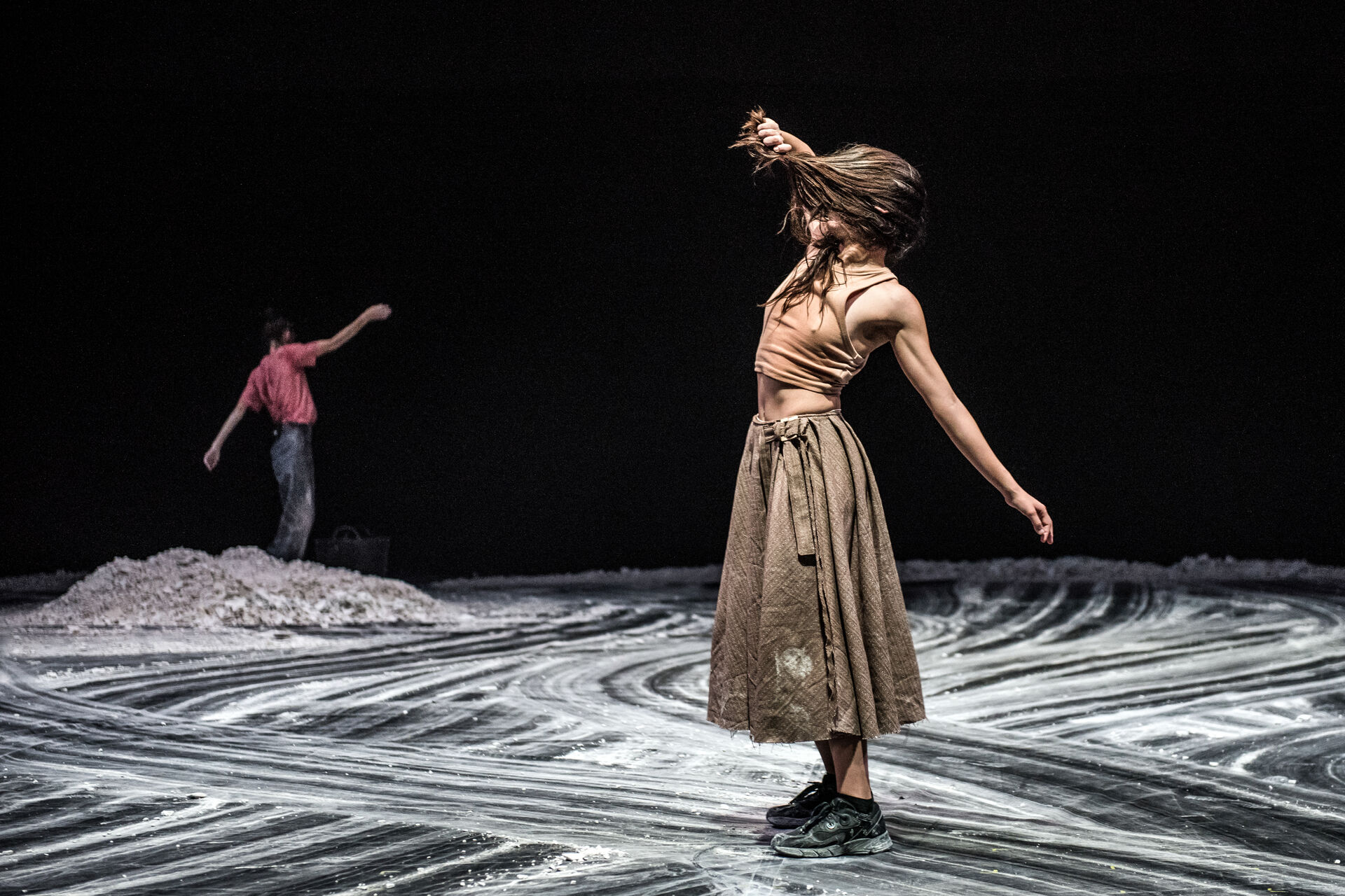 MINTATI: a dancer in a beige tank-top and brown skirt with hair flying mid-spin on a dark stage with sand-coloured swirling patterns under her feet; a second dancer in red is visible in the background. Athens Epidaurus Festival 2024, photo by Karol Jarek.