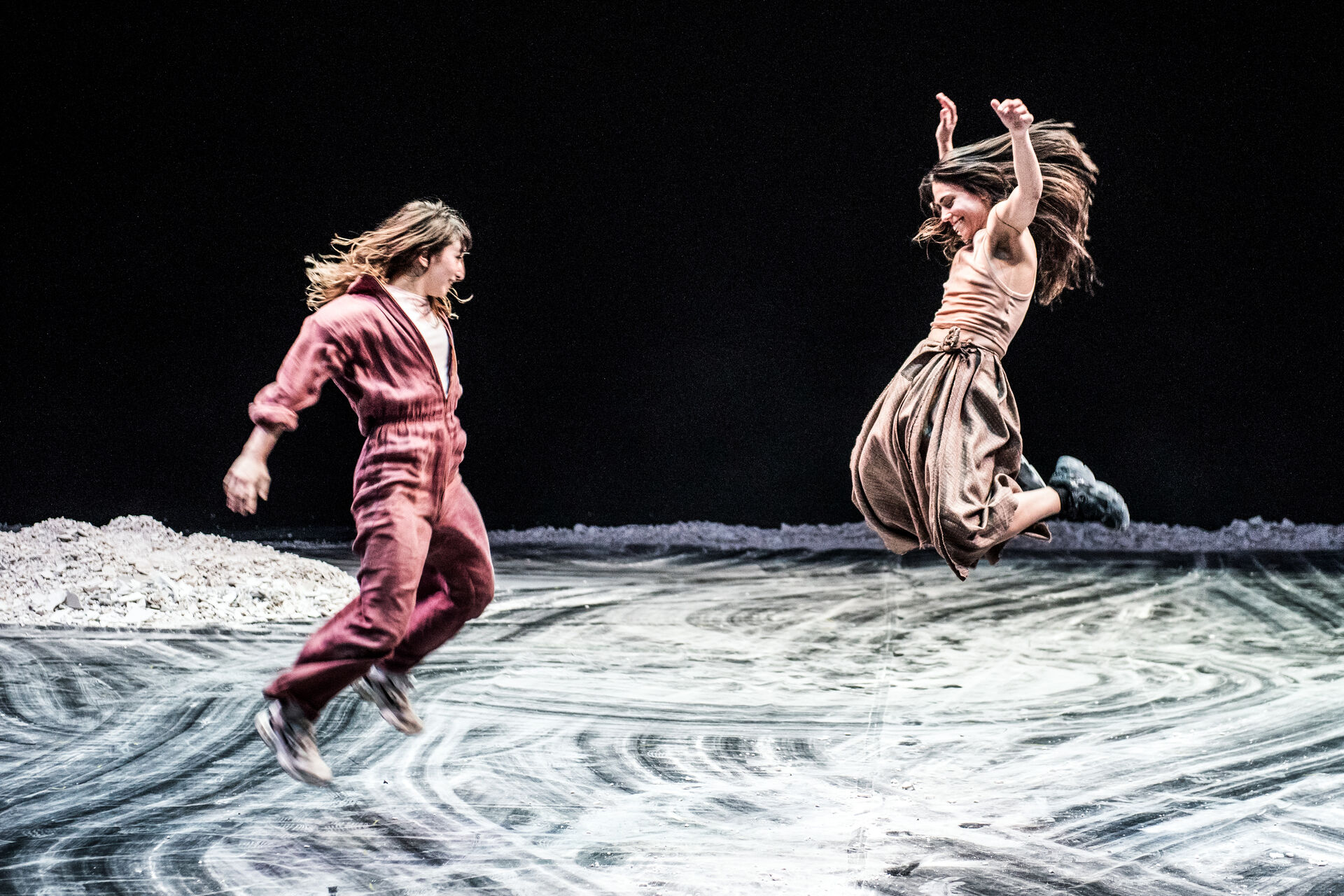 MINTATI: two dancers — one in a red jumpsuit, one in beige with a brown skirt — leap mid-air over swirling sand-pattern flooring, hair and arms thrown upward. Photo by Karol Jarek.