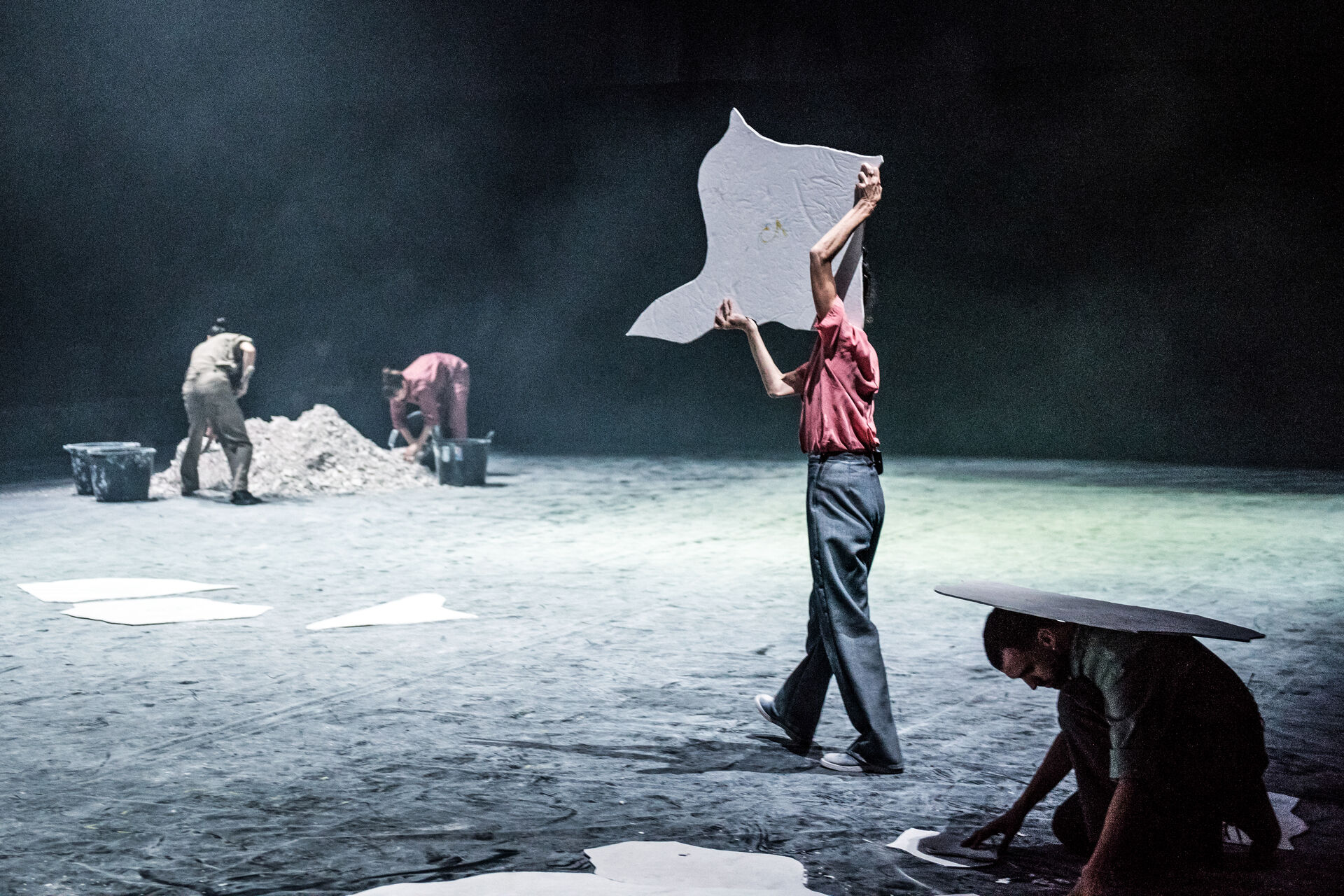 MINTATI: a dancer in a red shirt and jeans holds up a large irregular white sheet like a sail, while two performers behind work over a pile of earth and a third crouches under a dark angular shape; haze fills the dim stage. Photo by Karol Jarek.