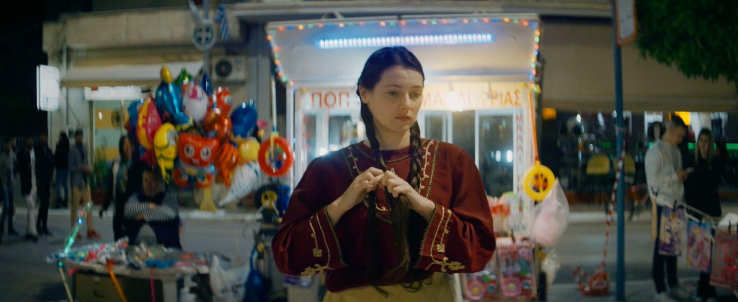 Mothers of Daughters still: a young woman in a red traditional-pattern dress stands at night in front of a kiosk lined with brightly coloured candies and balloons.