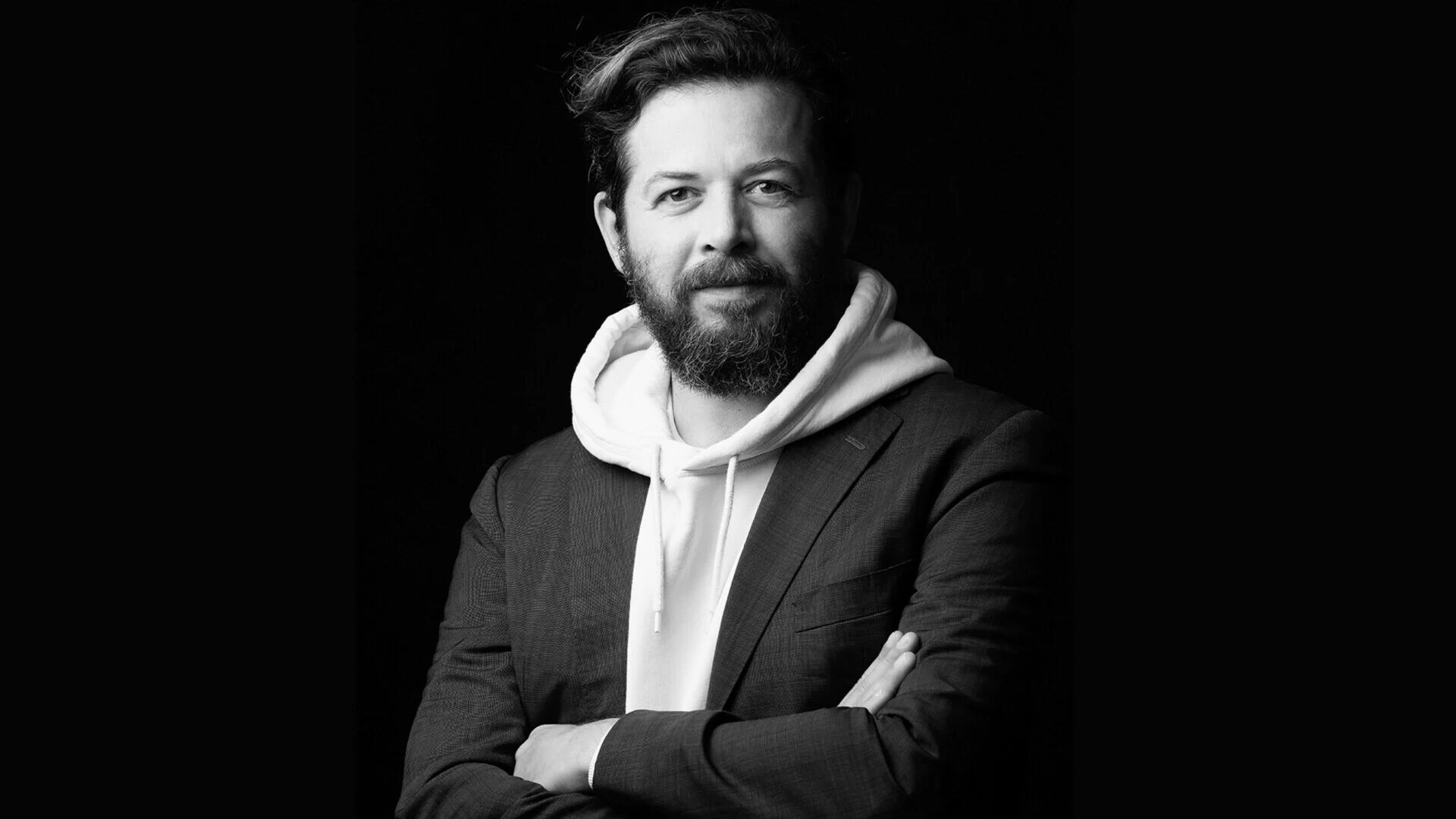 Black-and-white portrait of director Syllas Tzoumerkas: arms folded, wearing a hooded sweatshirt under a blazer, against a black background.