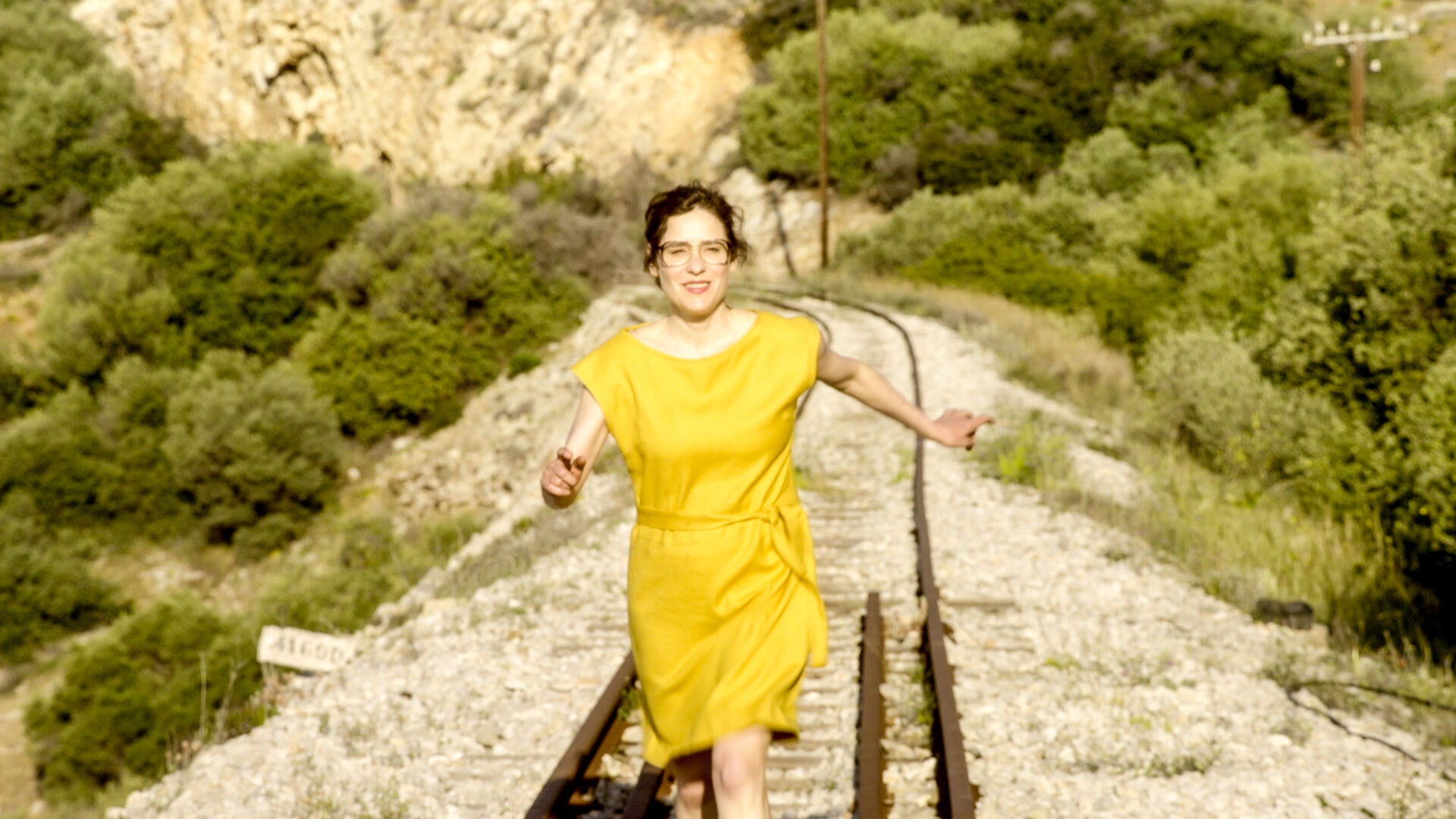 My Mother is a Saint still: actress Angeliki Papoulia in a yellow dress runs forward along sunlit railway tracks between green hillsides.