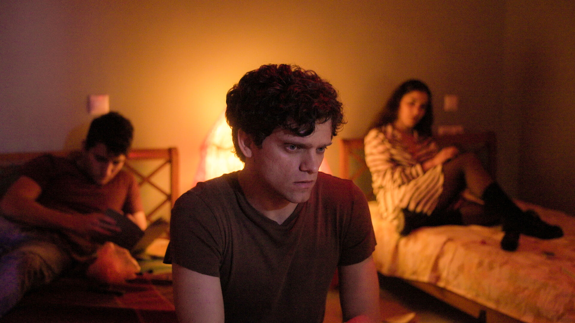My Mother is a Saint still: actor Jacopo Olmo Antinori in a brown t-shirt seated in a warmly lit room with an intent expression; two other people are visible on beds in the background.