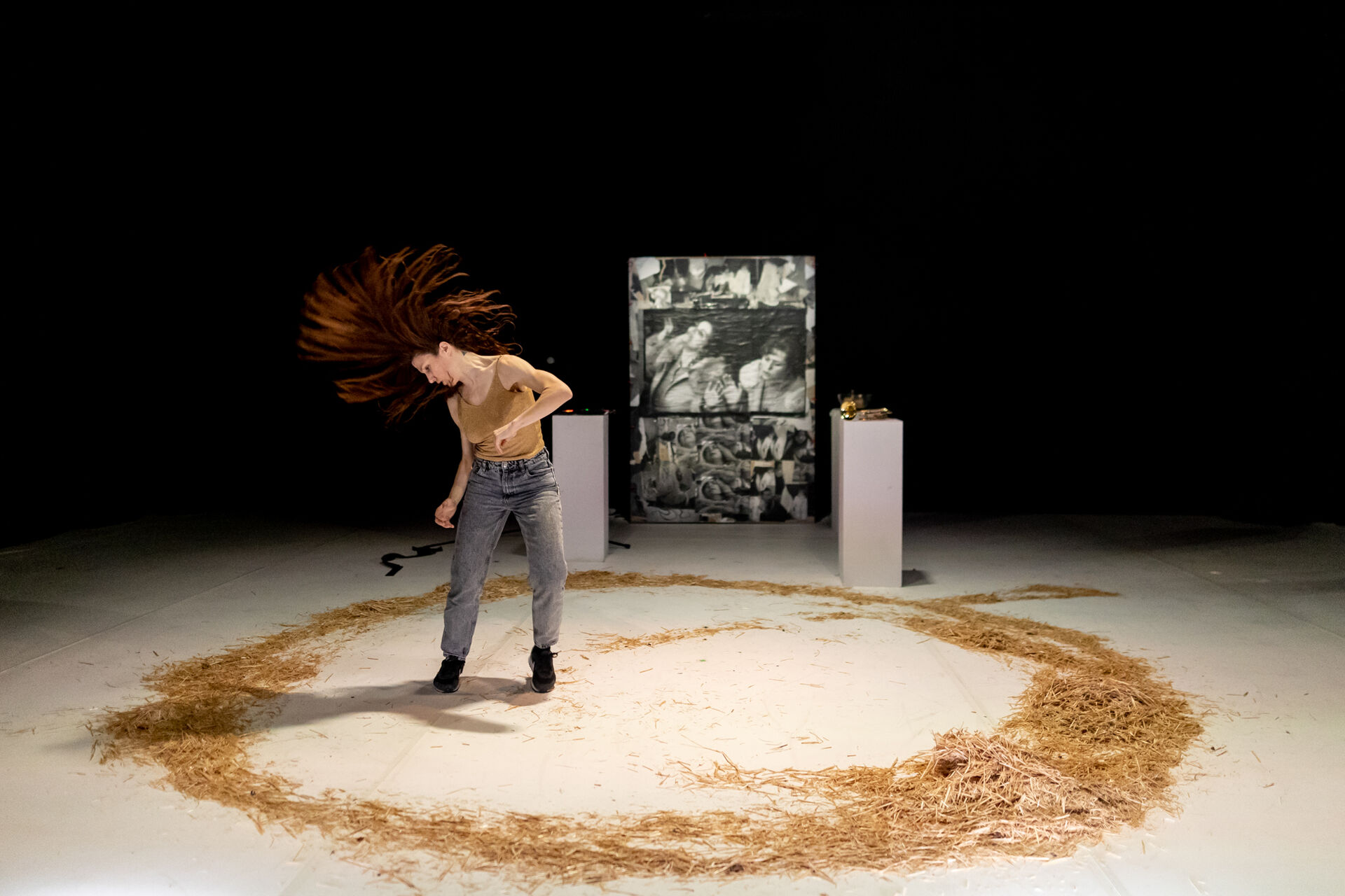 To Be Possessed: Chara Kotsali mid-spin with hair sweeping across her face on a white floor inscribed with a circle of dry straw; a dark painted panel rests on a plinth in the background.