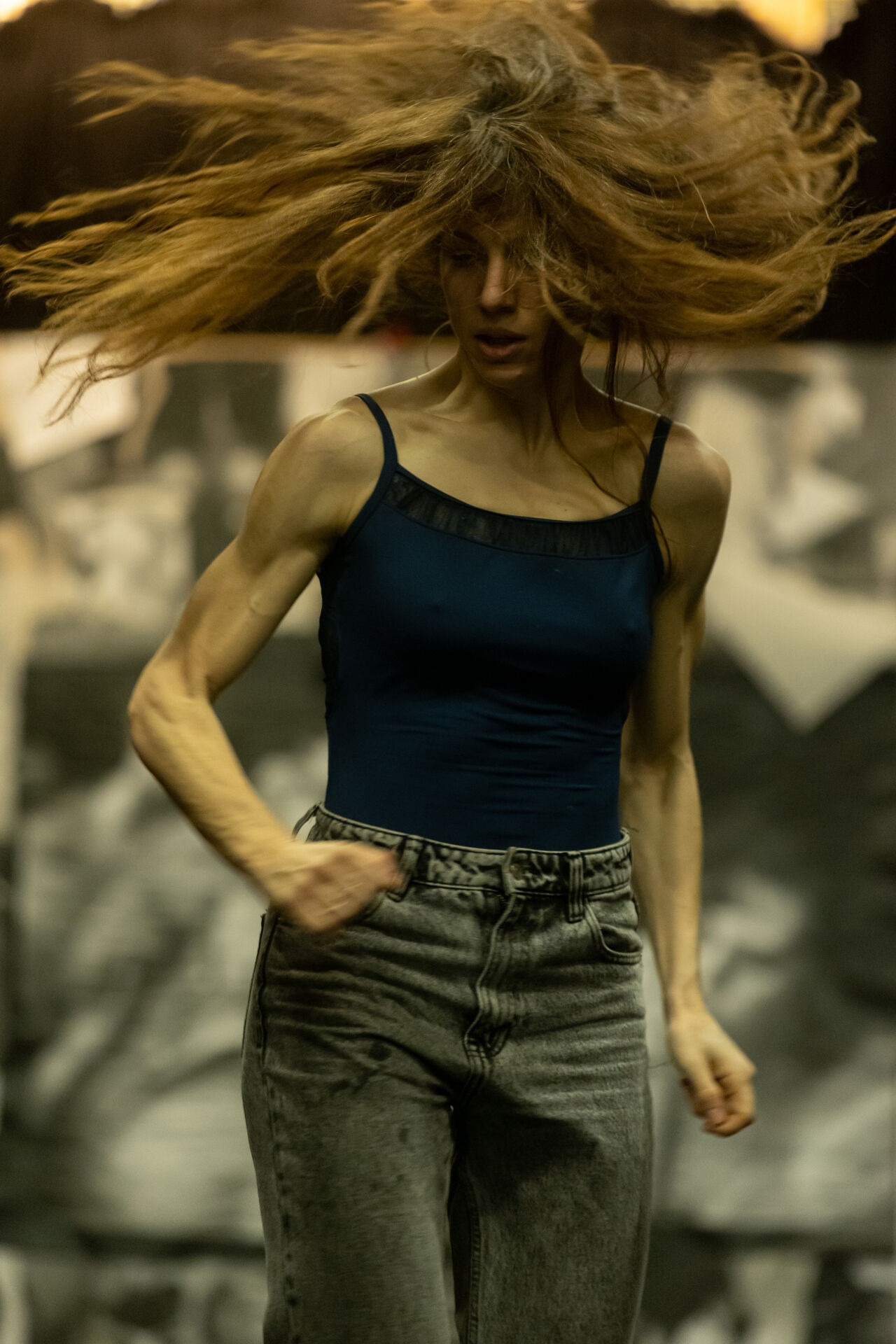 To Be Possessed: close-up of Chara Kotsali in a navy tank-top and grey jeans, hair flying upward in motion blur against a backdrop of black-and-white archival imagery.